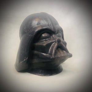 May include: A dark bronze-colored bust of Darth Vader, the iconic Star Wars character. The sculpture features the character's helmet and mask, with detailed facial features and a textured surface. The bust is a collectible item.