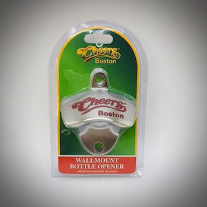 May include: A wall-mount bottle opener in a clear plastic package. The opener is silver with the words "Cheers Boston" in red and gold. The packaging has a green and yellow background.