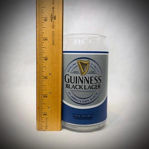 Może przedstawiać: Przezroczysty szklany kufel w kształcie puszki Guinness Black Lager z srebrno-niebieską etykietą. Etykieta zawiera logo harfy Guinness i tekst. Drewniana linijka mierzy około 15 cm.