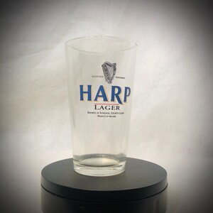 Puede incluir: Vaso de pinta transparente con el logotipo de Harp Lager en azul y dorado. El logotipo incluye un s&iacute;mbolo de arpa y el texto "Harp Lager", con texto debajo que indica la ubicaci&oacute;n de la cervecer&iacute;a en Dundalk, condado de Louth, Irlanda.