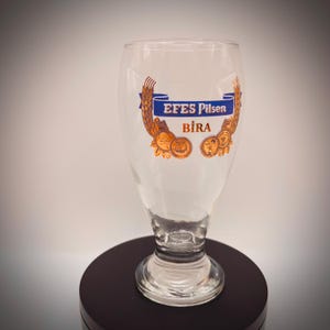 May include: Clear glass beer glass with a flared top and a rounded base. The glass features the text "EFES Pilsen Bira" in blue and orange, with wheat and coin-like designs. The glass is on a dark surface.
