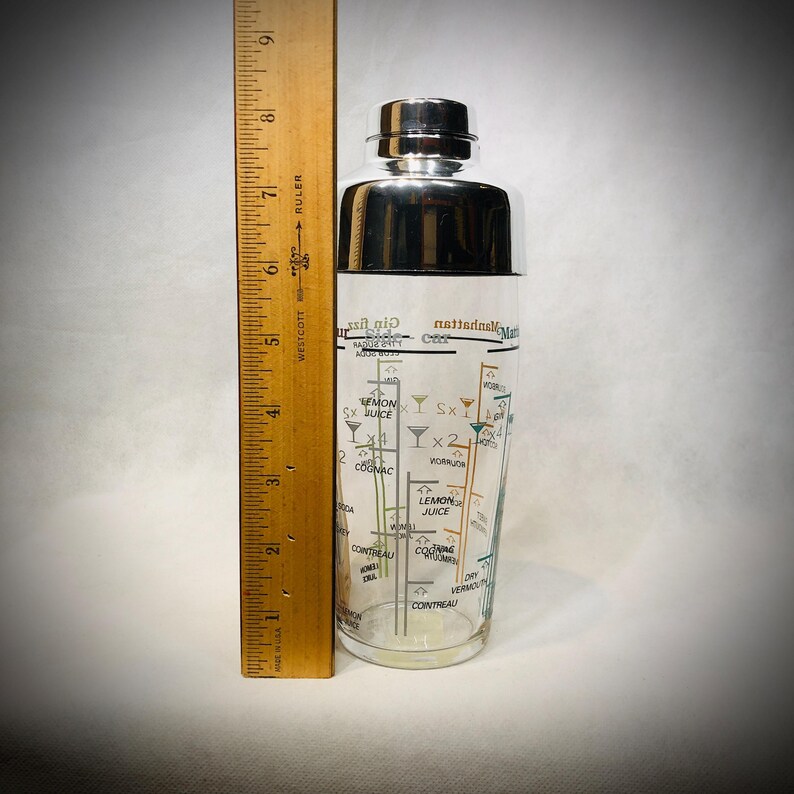Puede incluir: Una coctelera vintage con tapa de acero inoxidable y cuerpo de vidrio transparente. El vidrio est&aacute; impreso con recetas de c&oacute;cteles y medidas. Una regla de madera est&aacute; al lado de la coctelera.