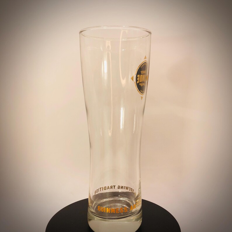 K&ouml;nnte beinhalten: Ein klares, hohes Bierglas mit leicht ausgestellter Form. Das Glas hat ein schwarzes und goldenes kreisf&ouml;rmiges Logo und goldenen Text mit der Aufschrift "Guinness". Das Glas ist leer und steht auf einer schwarzen Oberfl&auml;che.