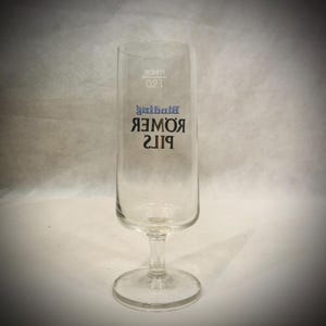 May include: A clear Pilsner glass with a stem and a wide base. The glass has the words "Romer Pils" printed on it in black and blue, with the word "Binibni" above it. The glass also has the measurement "0.2l" printed on it.