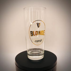 May include: A clear pint glass with the Guinness Blonde American Lager logo. The logo is a gold oval with the Guinness harp symbol and the words "BLONDE" and "AMERICAN LAGER" in gold lettering. The glass is on a black surface.