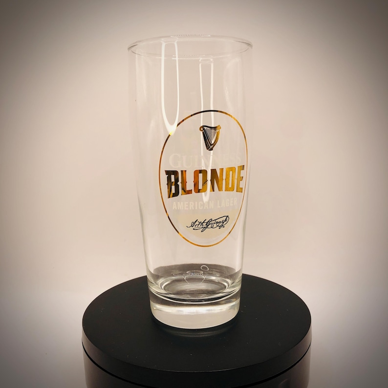 May include: A clear pint glass with the Guinness Blonde American Lager logo. The logo is a gold oval with the Guinness harp symbol and the words "BLONDE" and "AMERICAN LAGER" in gold lettering. The glass is on a black surface.