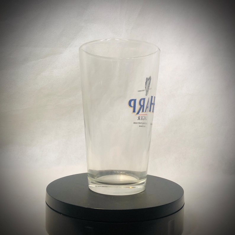 Puede incluir: Un vaso de pinta transparente con la palabra "Harp" en letras azules y un logotipo de arpa. El vaso est&aacute; vac&iacute;o y se encuentra sobre un soporte giratorio negro. El fondo es un degradado de blanco y gris.