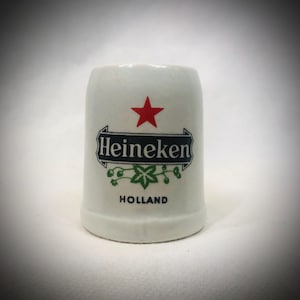 May include: A white ceramic shot glass featuring the Heineken logo with a red star, green accents, and the word "HOLLAND". The shot glass has a cylindrical shape.