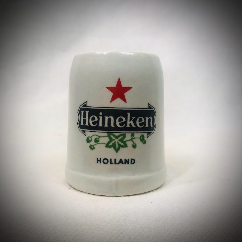 May include: A white ceramic shot glass featuring the Heineken logo with a red star, green accents, and the word "HOLLAND". The shot glass has a cylindrical shape.