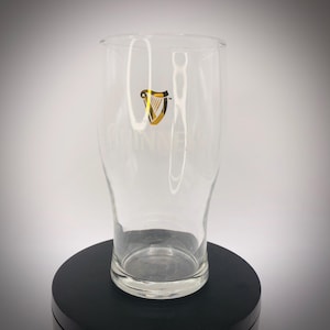 May include: A clear pint glass with the Guinness logo in gold and black. The glass has a curved shape and a wide base, with the Guinness name printed vertically. The glass is empty and sits on a black surface.
