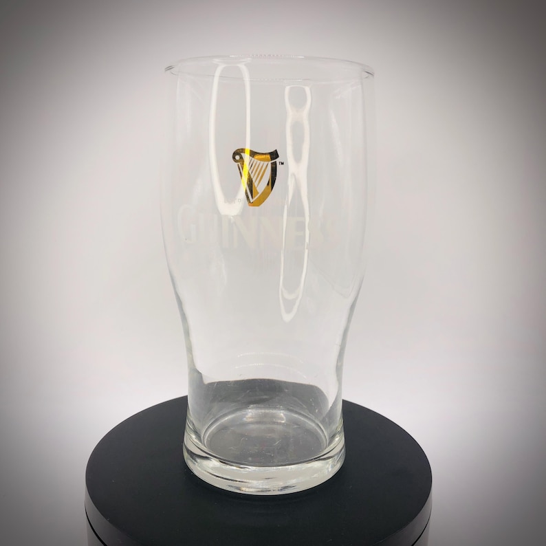 May include: A clear pint glass with the Guinness logo in gold and black. The glass has a curved shape and a wide base, with the Guinness name printed vertically. The glass is empty and sits on a black surface.
