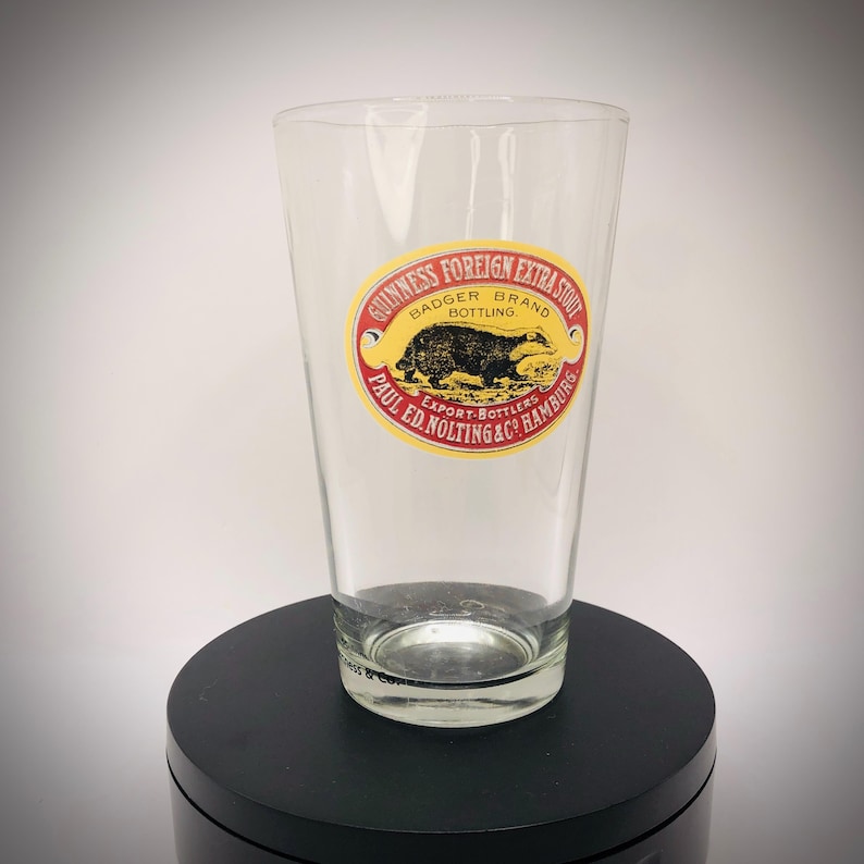 K&ouml;nnte beinhalten: Ein klares Pintglas mit einem Guinness Foreign Extra Stout Badger Brand Etikett. Das ovale Etikett hat roten, gelben und schwarzen Text und eine Dachs-Illustration. Das Glas ist leer und steht auf einer schwarzen Oberfl&auml;che.