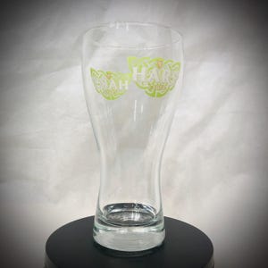 May include: A clear glass beer pint glass with a flared top and a sturdy base. The glass features green lettering that reads "HARP LAGER" in a Celtic-inspired design. The glass is empty and sits on a black surface.