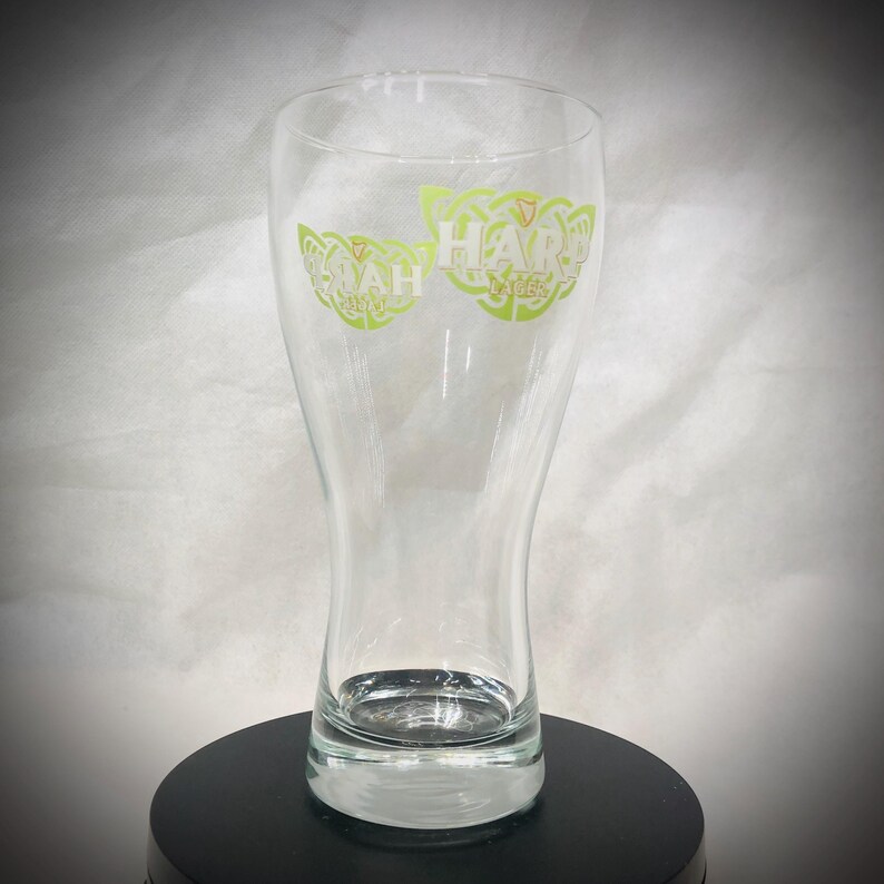 May include: A clear glass beer pint glass with a flared top and a sturdy base. The glass features green lettering that reads "HARP LAGER" in a Celtic-inspired design. The glass is empty and sits on a black surface.