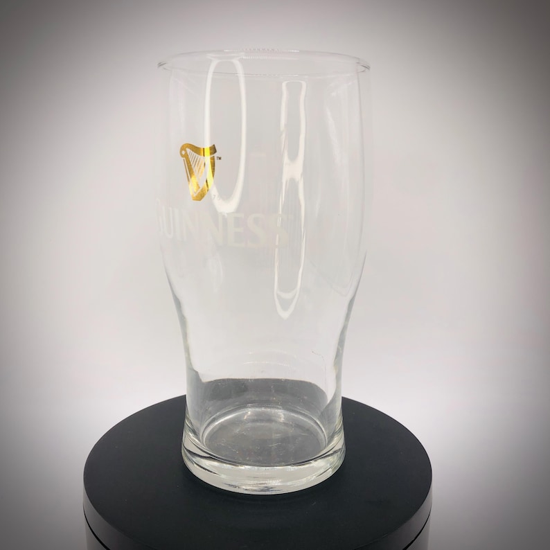 May include: A clear pint glass with the Guinness logo, featuring a gold harp, is displayed. The word "GUINNESS" is printed on the glass. The glass is on a black surface.