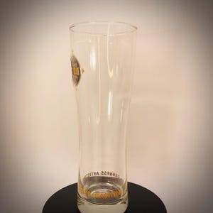 K&ouml;nnte beinhalten: Ein hohes, klares Bierglas mit leicht geschwungener Form. Das Glas hat ein Logo an der Seite und Text am Boden. Das Logo ist ein schwarzer Kreis mit goldenem Text. Der Text am Boden lautet "Guinness Tradition".