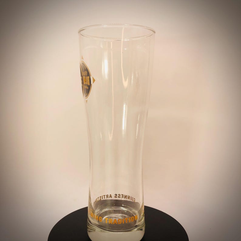 K&ouml;nnte beinhalten: Ein hohes, klares Bierglas mit leicht geschwungener Form. Das Glas hat ein Logo an der Seite und Text am Boden. Das Logo ist ein schwarzer Kreis mit goldenem Text. Der Text am Boden lautet "Guinness Tradition".