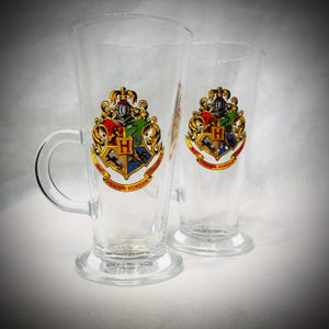 May include: Two clear glass mugs featuring the Hogwarts crest. The crest displays a shield divided into four sections, each representing a Hogwarts house, with the motto "Draco Dormiens Nunquam Titillandus". One mug has a handle.