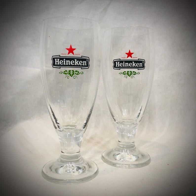May include: Two clear glass beer glasses with a stemmed base, each featuring the Heineken logo printed in black and red with a small green leaf graphic underneath. The glasses are transparent and have a curved, tapered shape for serving beer.