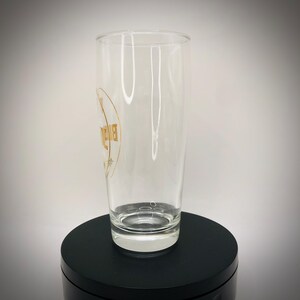 May include: A clear, tall glass with a gold logo on the side. The glass is cylindrical and sits on a black surface. The logo appears to be a brand name or emblem.