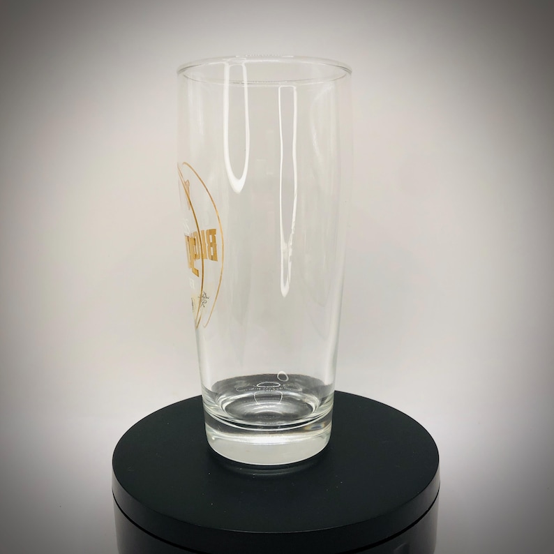 May include: A clear, tall glass with a gold logo on the side. The glass is cylindrical and sits on a black surface. The logo appears to be a brand name or emblem.
