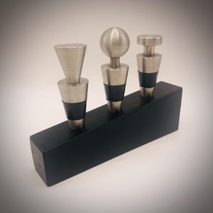 May include: Three wine bottle stoppers with brushed metal tops and black rubber seals are displayed on a black wooden base. The stoppers have different top designs: a cone, a sphere, and a flat disc.