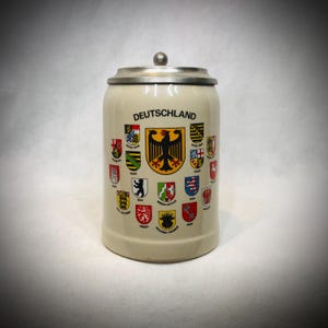 May include: A cream-coloured ceramic beer stein with a metal lid and a central black eagle emblem. The word DEUTSCHLAND is printed above the eagle, surrounded by various colourful regional coat of arms shields representing German states.
