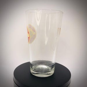K&ouml;nnte beinhalten: Ein klares Pintglas mit einem Logo an der Seite. Das Glas hat eine leicht konische Form und einen dicken Boden. Das Logo ist ein kreisf&ouml;rmiges Design mit roten, goldenen und wei&szlig;en Elementen. Das Glas ist leer und steht auf einer schwarzen Oberfl&auml;che.