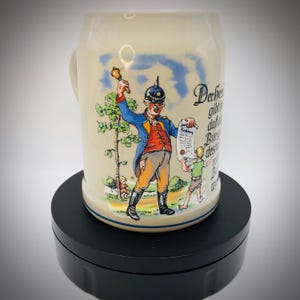 May include: Cream ceramic beer stein with a handle, decorated with a colourful illustration of a man in military uniform ringing a bell and holding a scroll. The stein has a blue rim and German text.