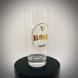 May include: A clear pint glass with a gold and white logo that reads "Guinness Blonde American Lager." The glass is cylindrical with a slightly flared top and a thick base. The logo includes a harp symbol.