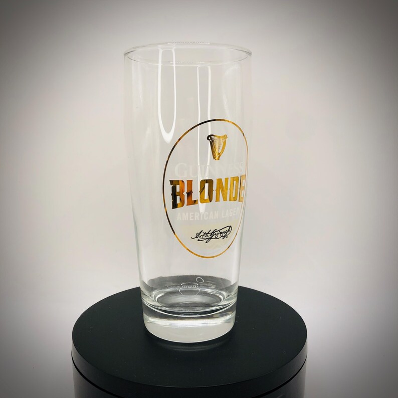 May include: A clear pint glass with a gold and white logo that reads "Guinness Blonde American Lager." The glass is cylindrical with a slightly flared top and a thick base. The logo includes a harp symbol.