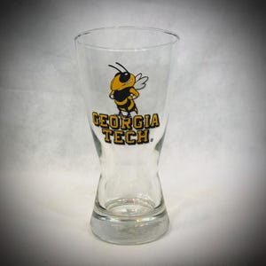 May include: Clear glass pint glass featuring a yellow and black cartoon bee mascot and the words "GEORGIA TECH". The glass has a flared top and a narrow waist. The glass is approximately 15 cm tall.