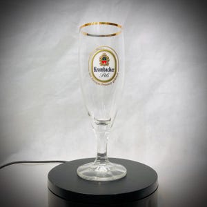 Puede incluir: Una copa de cerveza Pilsner transparente con borde dorado y un logotipo que dice "Krombacher Pils" en negro y dorado. La copa está sobre una base circular negra.