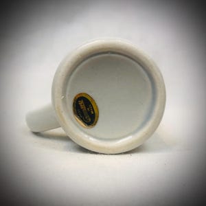 May include: A white ceramic mug with a handle, viewed from the bottom. A gold and black sticker is affixed to the base, with text that is difficult to read. The mug is on a white surface.