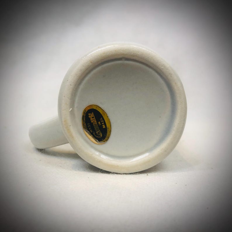 May include: A white ceramic mug with a handle, viewed from the bottom. A gold and black sticker is affixed to the base, with text that is difficult to read. The mug is on a white surface.