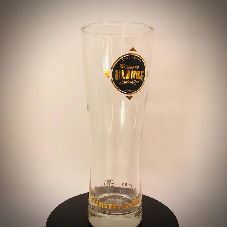 K&ouml;nnte beinhalten: Ein klares, hohes Bierglas mit geschwungener Form. Das Glas hat ein gold-schwarzes Logo mit der Aufschrift "Guinness Blonde American Lager Series". Der Boden des Glases tr&auml;gt den goldenen Schriftzug "Dentistry & Bar".