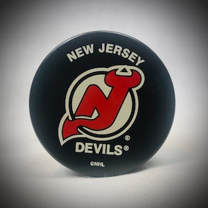 Könnte beinhalten: Ein schwarzer Hockeypuck mit dem Logo der New Jersey Devils. Das Logo zeigt ein rotes "N" mit stilisiertem Schwanz, weiß umrandet, und die Worte "NEW JERSEY" und "DEVILS" in weißer Schrift. Das NHL-Logo ist ebenfalls zu sehen.