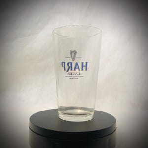 Puede incluir: Un vaso de pinta transparente con la palabra "HARP" en azul y un logotipo de arpa. El vaso tambi&eacute;n tiene la palabra "LAGER" impresa. El vaso est&aacute; sobre una superficie negra.