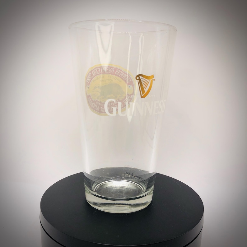 K&ouml;nnte beinhalten: Ein klares Pintglas mit dem Guinness-Logo. Das Glas hat eine goldene Harfe und den Namen Guinness in Wei&szlig; aufgedruckt. Ein kreisf&ouml;rmiger Aufkleber mit Text und einem B&auml;rengrafik ist ebenfalls sichtbar.