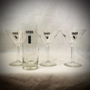 Puede incluir: Cuatro copas de cóctel transparentes. Dos copas de martini están etiquetadas "SHAKEN" con un gráfico de coctelera, una está etiquetada "STIRRED" con un gráfico de cóctel, y un vaso alto está etiquetado "CRUSHED" con un gráfico de licuadora. Todo el texto y los gráficos son negros.