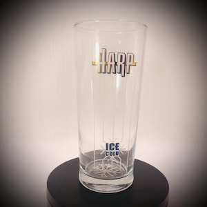 May include: A clear glass with the word "Harp" in gold and black lettering. The glass has vertical lines and the words "Ice Cold" in blue. The glass is on a black surface.