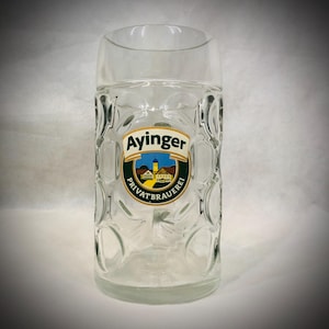 Könnte beinhalten: Ein klarer Bierkrug mit Henkel und einem strukturierten, geriffelten Design. Der Krug hat ein farbenfrohes Logo mit dem Text "Ayinger Privatbrauerei". Das Logo zeigt ein Gebäude und einen blauen Himmel.