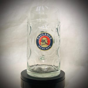Può includere: Un boccale da birra trasparente con un esterno strutturato. Il boccale presenta un logo circolare con le parole "PAULANER MÜNCHEN" e l'immagine del profilo di un uomo. Il boccale è progettato per contenere una grande quantità di birra.