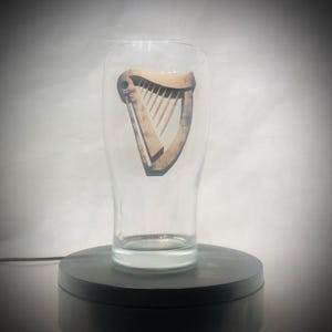 Puede incluir: Un vaso de pinta transparente con el logotipo del arpa. El arpa es de color marr&oacute;n claro con un contorno oscuro. El vaso tiene forma cil&iacute;ndrica con una parte superior ligeramente ensanchada. El vaso est&aacute; sobre una superficie negra.