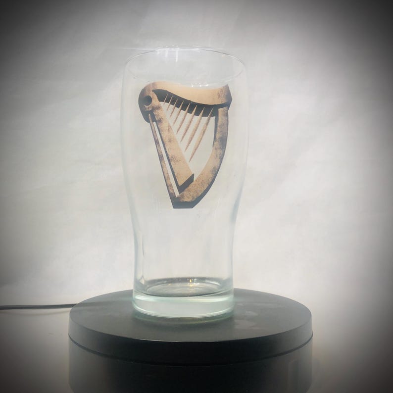 Puede incluir: Un vaso de pinta transparente con el logotipo del arpa. El arpa es de color marr&oacute;n claro con un contorno oscuro. El vaso tiene forma cil&iacute;ndrica con una parte superior ligeramente ensanchada. El vaso est&aacute; sobre una superficie negra.