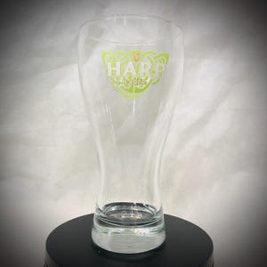 May include: A clear glass beer pilsner with a flared shape. The glass features the Harp Lager logo in green and white. The glass is empty and sits on a black surface.