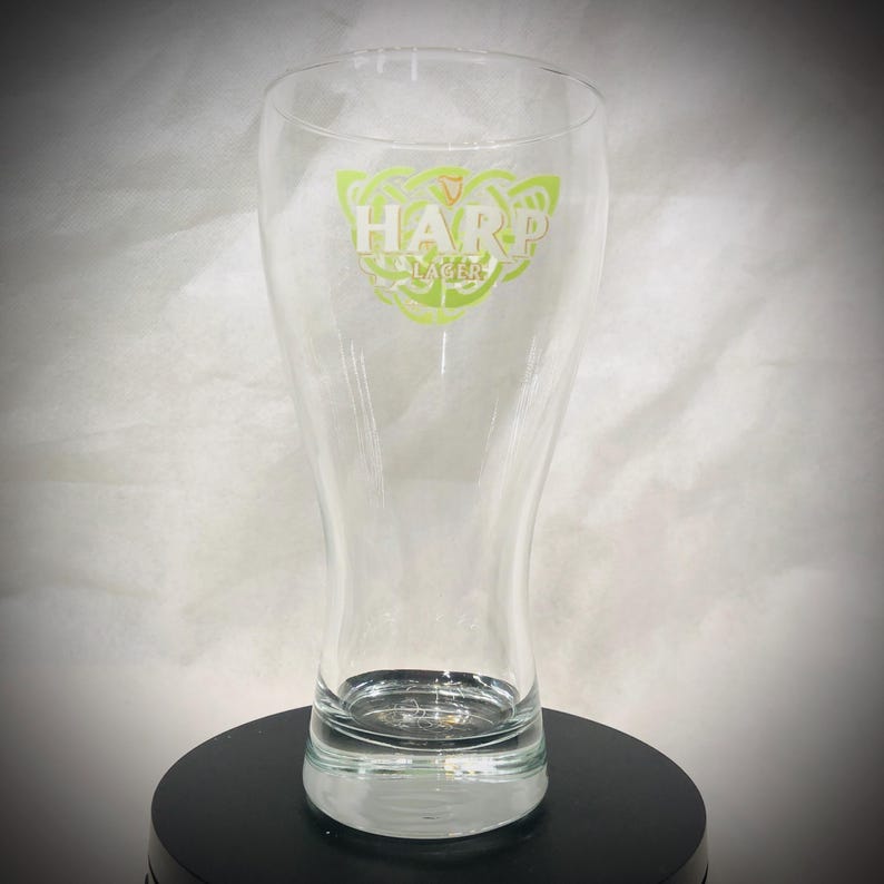 May include: A clear glass beer pilsner with a flared shape. The glass features the Harp Lager logo in green and white. The glass is empty and sits on a black surface.