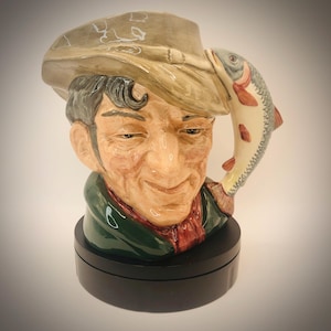 May include: A ceramic mug shaped like a smiling man wearing a hat, with a fish attached to the brim. The mug has a green collar and a red scarf. The fish is white with red and gray details. The mug sits on a black base.