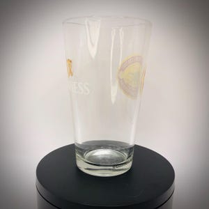 K&ouml;nnte beinhalten: Ein klares Pintglas mit dem Guinness-Logo und einem runden Aufkleber. Das Glas ist zylindrisch mit einem leicht ausgestellten oberen Rand. Das Guinness-Logo ist goldfarben und das Wort "Guinness" ist wei&szlig;. Es steht auf einer schwarzen Oberfl&auml;che.