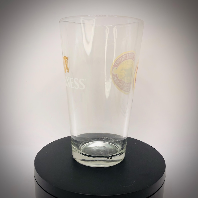 K&ouml;nnte beinhalten: Ein klares Pintglas mit dem Guinness-Logo und einem runden Aufkleber. Das Glas ist zylindrisch mit einem leicht ausgestellten oberen Rand. Das Guinness-Logo ist goldfarben und das Wort "Guinness" ist wei&szlig;. Es steht auf einer schwarzen Oberfl&auml;che.